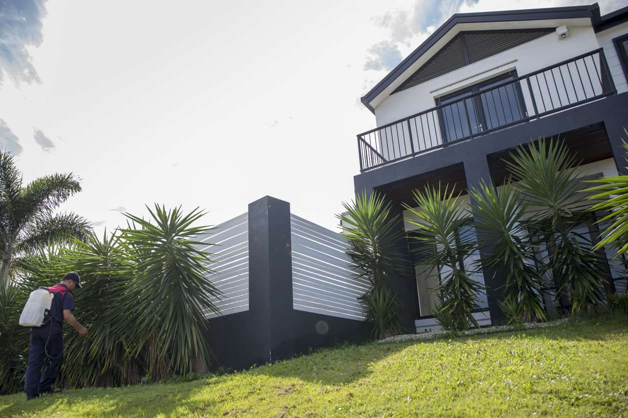 A pest control specialist spraying around the plants and fencing outside a large multistory home.